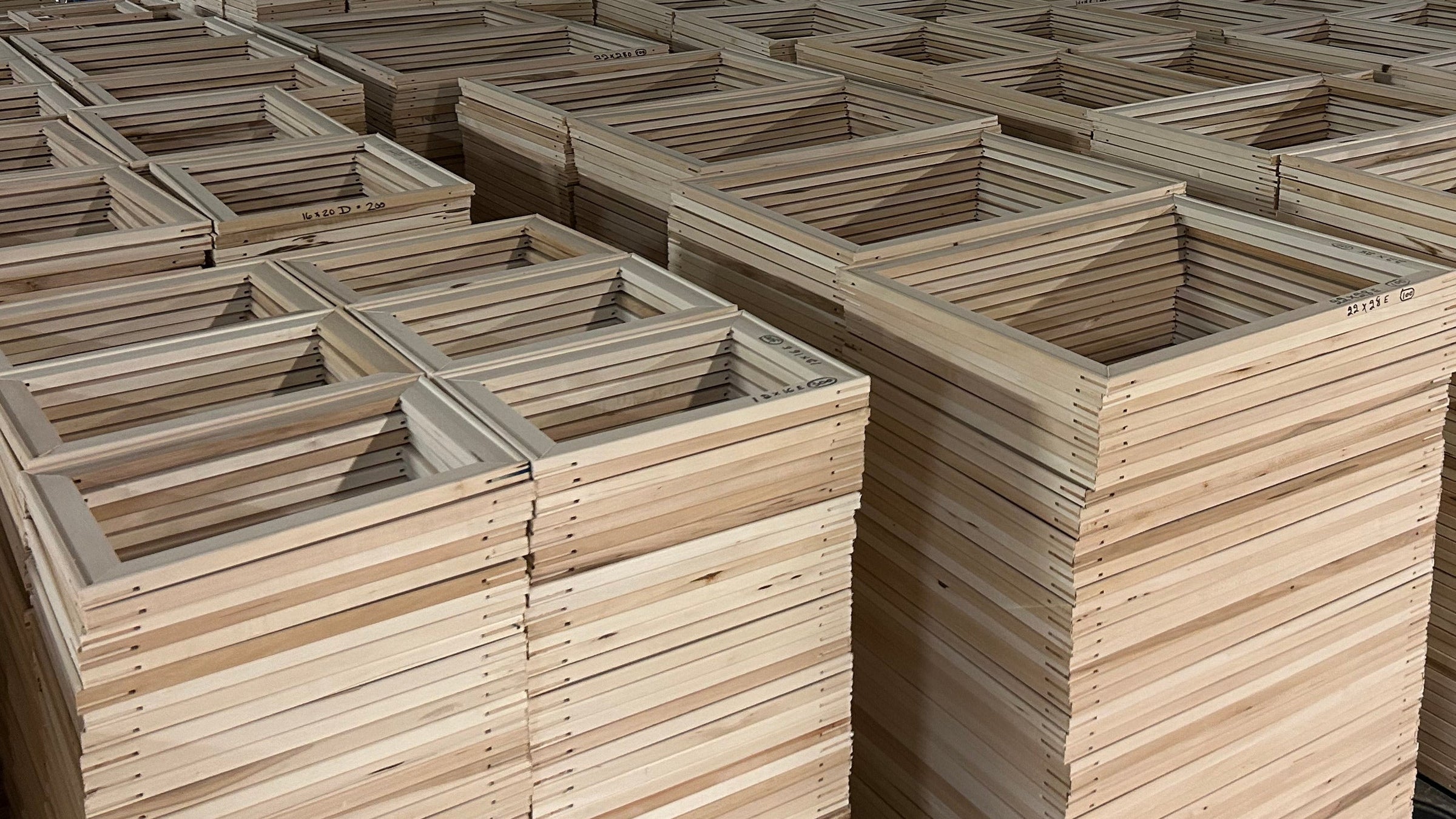 Large stacks of 63 series 3/4" stretcher bars wood frames on pallets arranged in rows inside a warehouse or production facility with metal roofing and industrial equipment visible in the background.