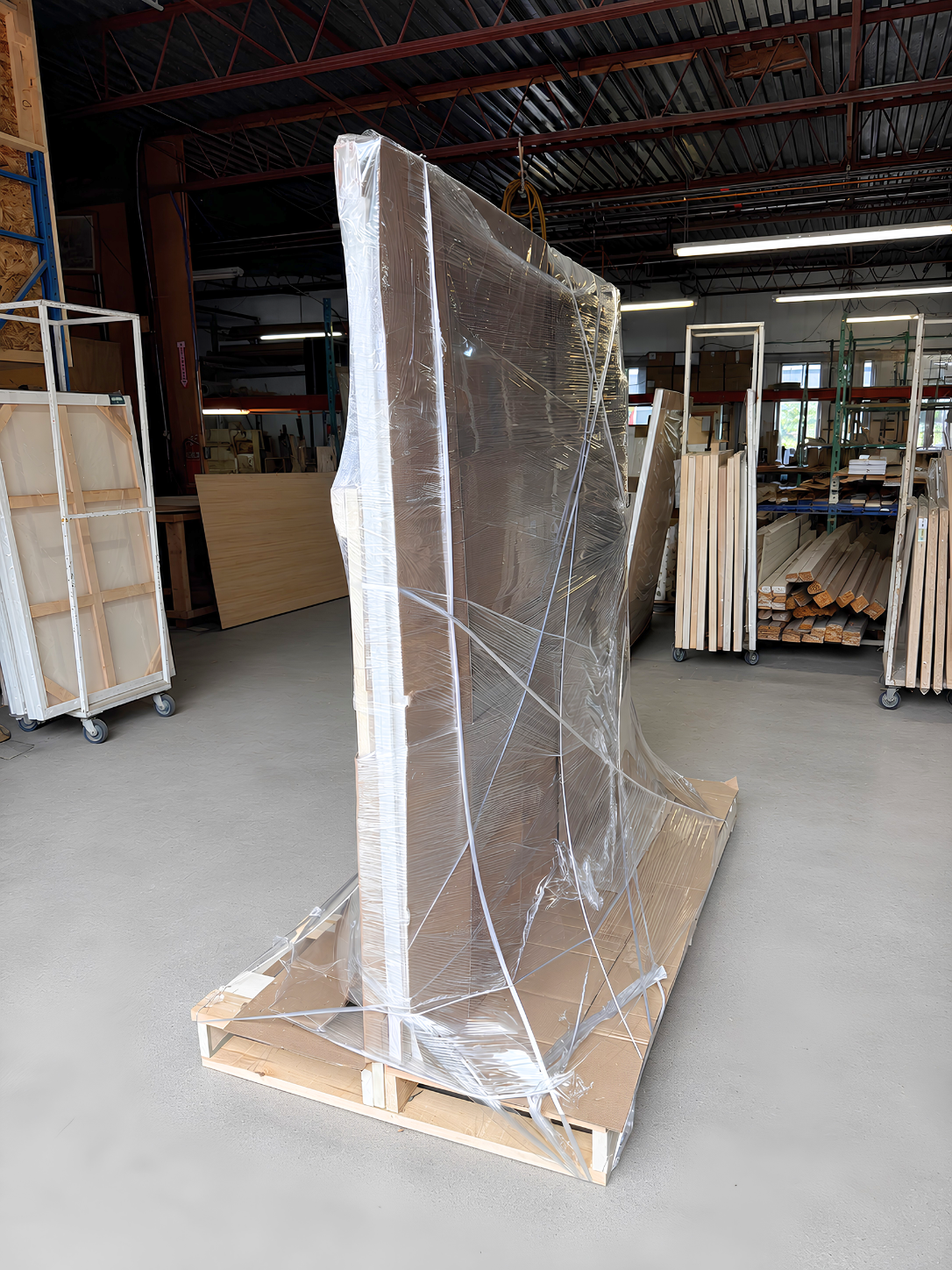 A large rectangular panel wrapped in clear plastic and secured on a wooden pallet inside the Apollon warehouse. Similar wrapped panels and stacks of wood are visible in the background.