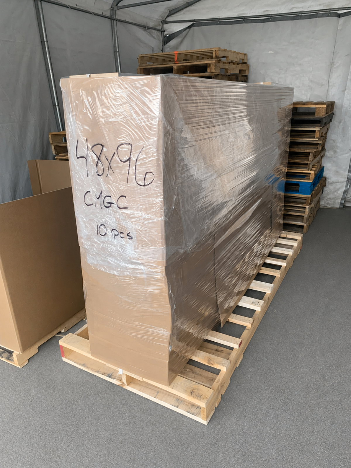A large, tall rectangular cardboard box wrapped in clear plastic film sits on a wooden pallet inside the Apollon storage area. Handwritten text on the side reads '48x96 CM/C 10 pcs'.