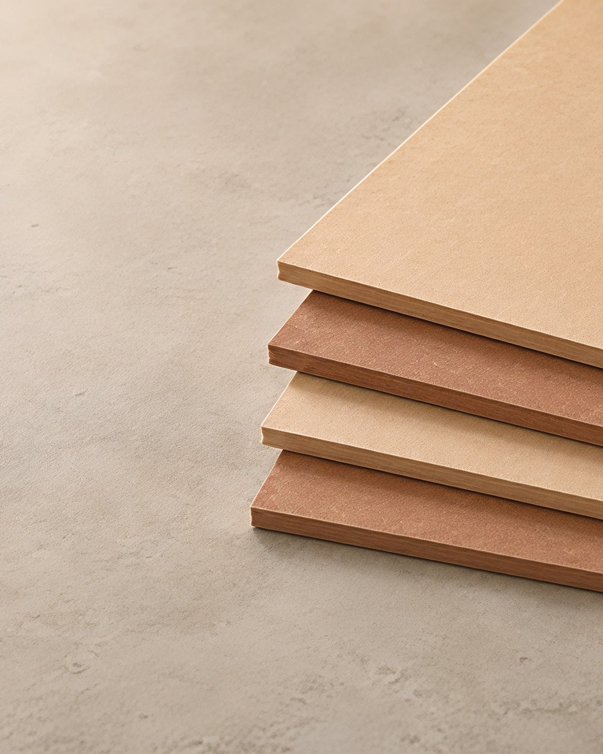 Quatre feuilles rectangulaires empilées de massonite dans des tons de brun foncé et clair, disposées sur une surface de béton gris clair texturée.